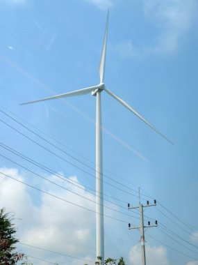 Enerji hatları ve yeşilliklerle dolu, yenilenebilir enerji ve sürdürülebilirliği sembolize eden parlak mavi gökyüzüne karşı yükselen bir rüzgar türbini..