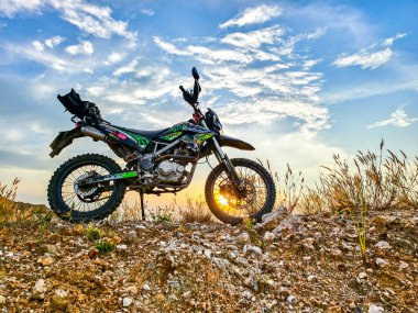 Kayalık zeminde gün batımı ve dramatik bulutlarla dinamik bir motosiklet görüntüsü..