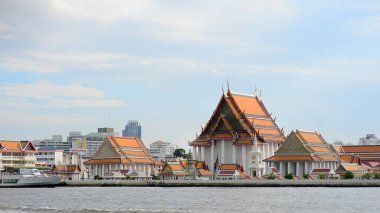 Nehir kenarındaki geleneksel Tayland tapınağının manzarası. Arka planda açık mavi bir gökyüzünün altında modern bir şehir silueti var..