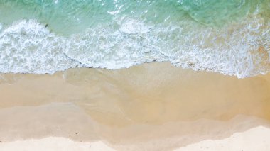 Havadan çekilen görüntüler kumlu bir kumsalın sakin güzelliğini yakalıyor. Yumuşak dalgalar açık bir gökyüzünün altında kıyıya vuruyor..