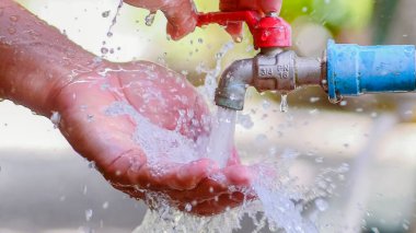 Eller kırmızı musluktan temiz su topluyor, açık hava ortamında tazeliği ve sürdürülebilirliği simgeliyor..