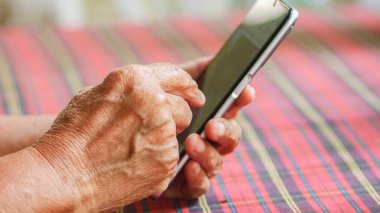 Yaşlılar arasında teknoloji kullanımını vurgulamak için canlı bir masa örtüsü üzerinde akıllı telefon kullanan yaşlı ellerin yakın çekimleri.