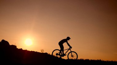 Bir bisikletçi çarpıcı bir günbatımı gökyüzüne karşı siluet, arka planda güneş batarken yokuş yukarı at sürüyor.