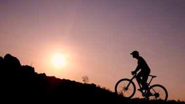 Bir bisikletçinin silueti gün batımında bir tepeye tırmanıyor, doğadaki macera ve sükunetin özünü yakalıyor..