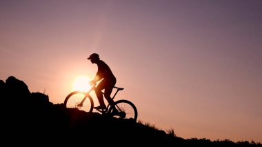 Göz kamaştırıcı bir gün batımına karşı silueti olan bir bisikletçi, parlak bir gökyüzü ve parlayan güneşle dağ bisikletine biniyor..