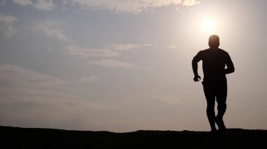 Gün batımında tepenin üstünde koşan bir adamın silüeti. Arka planda dramatik bir gökyüzü ve güneş var..