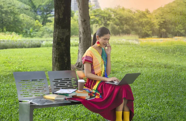 woman transgender in colorful rainbow dresses sitting on bench in the park, the lgbtq transgender is typing laptop computer and using smartphone. concept lgbtq lifestyle, lgbtq pride month