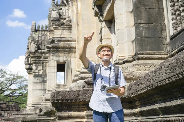 senior man with grey hair and beard,wearing hat,carry backpack and having camera, holding tablet in hand is pointing to something. concept ancient tourist, ecotourism, archeologist