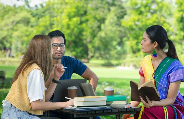 university students learning and doing research together on weekend in the park,group of friends sitting and discussing work, there are computer laptops, text books,coffee cups on table