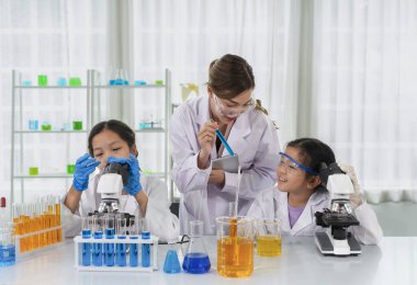 chemical laboratory staff as a mentor teaching kids in science classroom, young female hand holding tablet and other hand showing liquid tube and talking to a little kid 