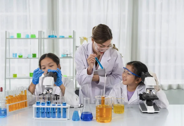 chemical laboratory staff as a mentor teaching kids in science classroom, young female hand holding tablet and other hand showing liquid tube and talking to a little kid