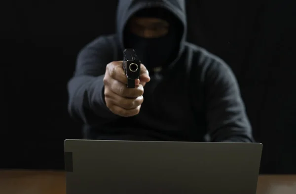 hacker in black hoodie with mask cover face poining a gun to camera, there is laptop on table,concept for financial cybercrime and fraud