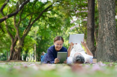 happy senior married couple spend their time relaxing in the park, they are lying on grasses, older adult man reading a book and a woman playing computer tablet