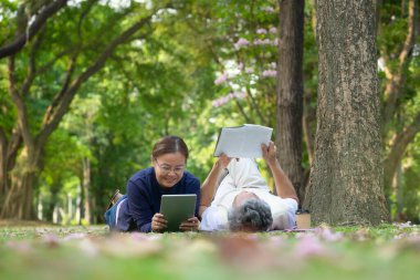 happy senior married couple spend their time relaxing in the park, they are lying on grasses, older adult man reading a book and a woman playing computer tablet