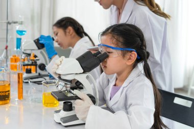 Mikroskop kullanmaya çalışan, fen bilimleri sınıfında kimya laboratuarı deneyi yapan bilim adamı üniformalı Asyalı küçük kız portresi. 