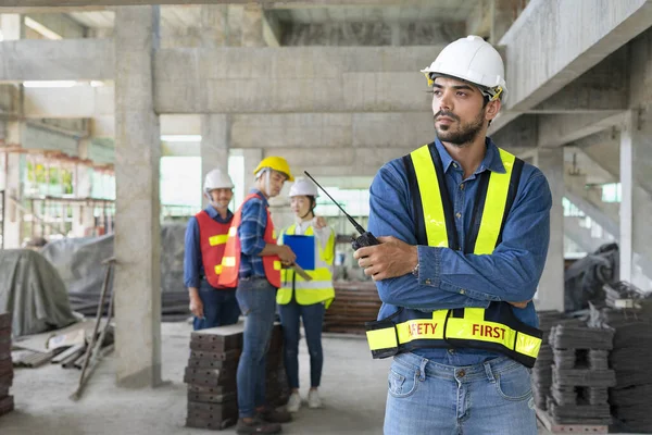 young confident engineer with arms crossed,holding walkie talkie, standing under construction building, background group of colleagues talking and bullying him. concept for management conflict in the workplace