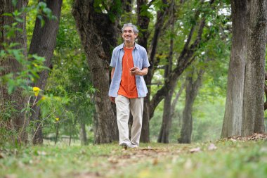 Gri saçlı, cep telefonu tutan, müzik dinlemek için kulaklık takan, gülümseyen, orman parkında yürüyen son sınıf öğrencisi. konsept yaşlı sağlık faydaları doğa, refah