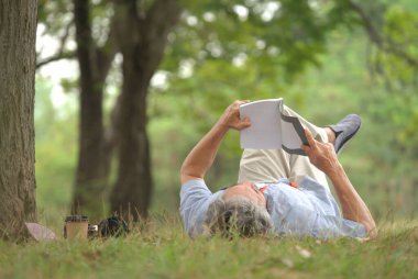Otların üzerinde yatan gri saçlı yaşlı adam, parkta gülümseyen ve kitap okuyan yaşlı adam, yaşlılar yaşam tarzı, hobiler, dinlenme, rahatlatıcı, doğada yaşlı sağlık faydaları, refah konsepti