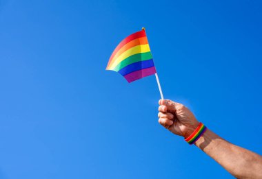 lgbt gökkuşağı bilekliği takan bir adamın eli havada lgbtq gökkuşağı bayrağı sallıyor mavi gökyüzü, LGBTQ + toplum eşitliği hareketi konsepti, lgbt mutlu gurur ayı