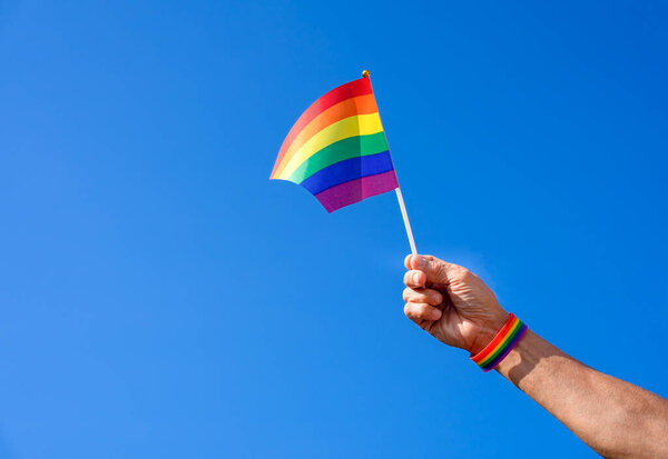 hand of a man wearing lgbt rainbow wristband waving lgbtq rainbow flag into the air background blue sky, concept for LGBTQ+ community equality movement, lgbt happy pride month