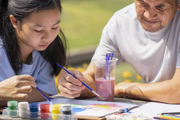 stock image asian teenage girl painting watercolors with her grandfather, happy family granddaughter and grandfather spend leisure time together in the garden, concept family lifestyle, relationship,the love of a family