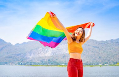 Genç Asyalı kadın elinde lgbt renkli gökkuşağı bayrağı, arka plan mavi gökyüzü, mutlu lezbiyen kadın lgbtq gay bayrağı sallıyor ya da çeşitlilik cinsiyetlerine saygı, homoseksüel, biseksüel, transseksüel, eşcinsel