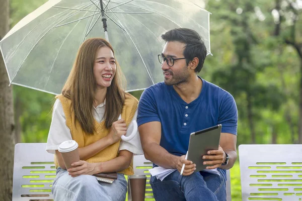 stock image romantic asian couple dating,studying or working in the garden on sunny day ,they raising an umbrella and talking together,young man and woman lifestyle sitting on bench in the park on weekend