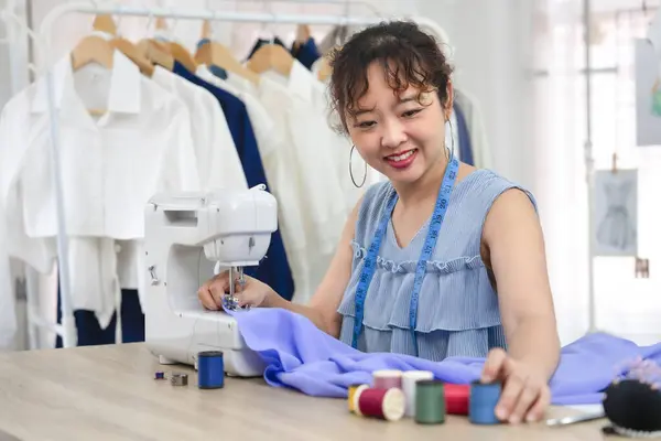 happy young asian female sitting with sewing machine,working, smiling experienced tailor is sewing clothes at fashion studio,concept of sewing,craft,tailoring,business,industry