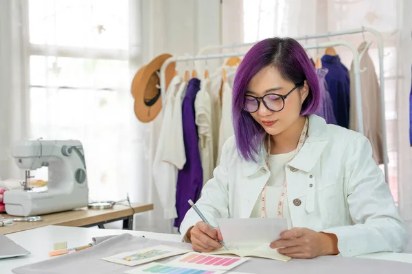 young cool female designer with purple color hair concentrate to sketching clothing design on table,a professional tailor working in fashion studio. concept clothing design,tailoring,business,industry