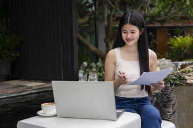 Genç ve çekici Asyalı bir kadın tatil boyunca tatil beldesindeki dinlenme yerinde dizüstü bilgisayar başında çalışıyor, dijital göçebe kavramı, özgürce seyahat ediyor, internette uzaktan çalışıyor. 