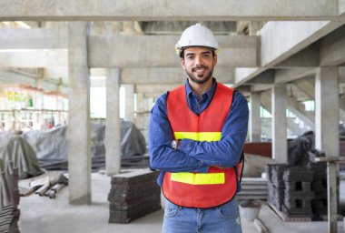 Güvenlik teçhizatı takan çekici genç erkek mühendis, kollarını kavuşturmuş, kendinden emin gülümsüyor, arka plan inşaat altındaki yeni bir binayı bulanıklaştırıyordu.