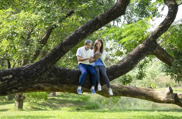 father and daughter enjoy relaxing in nature,sitting on a big tree,looking at tablet online together,concept for elderly people lifestyle,relaxing,wellbeing,family relationship,love of a family