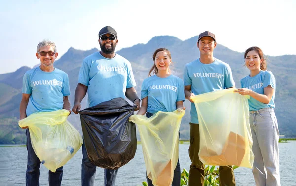 multiethnic people in blue volunteer T-shirt volunteers holding waste sorting plastic bag standing by the river,a diverse group of volunteers join together to cleanup the sand beach together