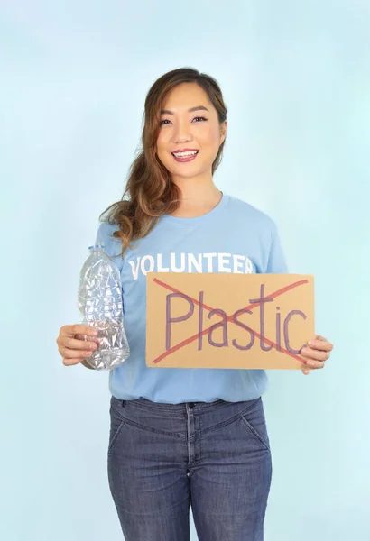female volunteer in blue T-shirt,hand holds used plastic bottle,other hand holds cardboard sign no plastic,on blue background,concept campaign to recycle used empty plastic bottles,stop global warming