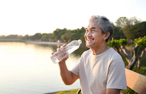 a smiling senior man resting by the pond in the public park,older adult male sitting and drinking water after workout,a healthy elderly man with grey hair and beard relaxing at sunset