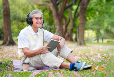 Müzik dinlemek için kulaklık takan, kitap tutan, el ele tutuşan gözlük takan, doğada gülümseyen Asyalı son sınıf öğrencisi yaşlı bir erkek doğa parkında hobi olarak vakit geçiriyor.