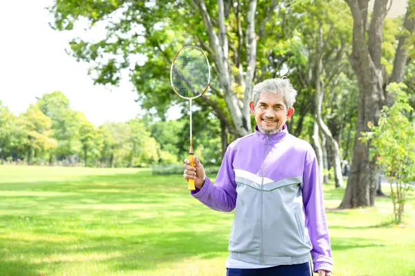 portrait asian senior man in sportswear having a badminton racket and looking at a camera,older adult male playing badminton outdoor in the park,concept elderly lifestyle,health care,exercising