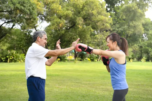 An elderly father and young adult daughter in sportswears are enjoying boxing in the summer park,concept of family lifestyle,family relationship,exercise,health care,health benefit in nature