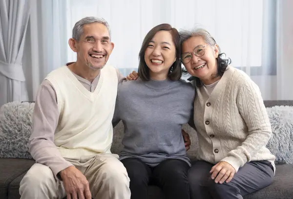 portrait happy asian family,older adult father and mother with young adult daughter sitting on couch,hugging together,smiling and looking at camera