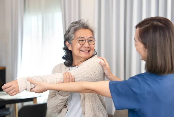 Olumlu duygular besleyen son sınıf öğrencisi kadın genç bir kadın hemşireden, Asyalı yaşlı bir yetişkinin rehabilitasyon kolunun evde gerilmesi, yaşlı yaşam tarzı, ev bakımı, evde sağlık hizmetleri kavramı.