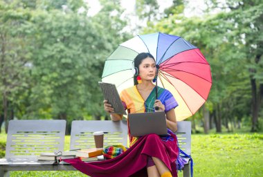 A transgender woman in colorful dresses and accessories wearing headphones,raising lgbt rainbow umbrella,sitting on bench,studying and relaxing in the park,concept of lgbtq community equality movement