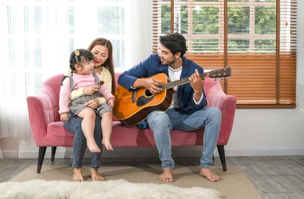 young interracial parent with a little daughter sitting on sofa playing guitar,singing a song,family spend leisure time together in the living room,concept of family,relationship,the love of a family