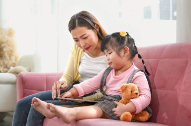 Genç Asyalı kadın çocuğuyla tablet kullanıyor. Annesi hafta sonları boş zamanını küçük kızıyla birlikte oturma odasında geçiriyor. 