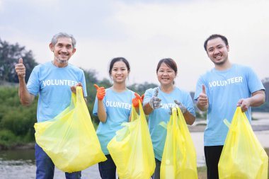 Bir grup aile gönüllüsü mavi tişörtle ayakta duruyor, ellerinde sarı plastik torbalar ve nehir kenarında başparmak hareketlerini yapıyorlar.
