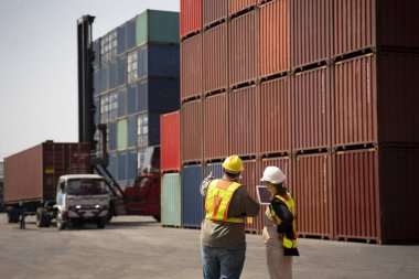 Kafkasyalı hamile kadın mühendis elinde dijital tabletle konteynır terminalinde çalışan, başparmak kaldıran, kargo, nakliye, lojistik işçisi şişman bir adamla konuşuyor.