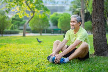 Spor giyimli yaşlı bir erkek, bir ağacın altında oturuyor, şehir parkında bacaklarını açıyor, olgun bir adam sabah güneşinde yeşil doğada kaygısız bir duygu yaşıyor, yaşlı yaşam tarzı, refah, refah kavramı...