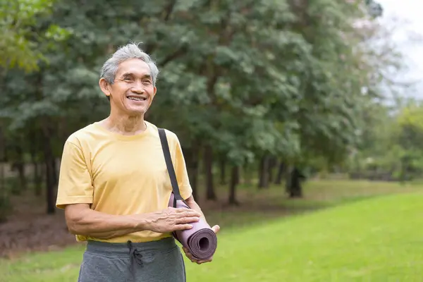 portrait asian senior man with yoga mat standing in the summer park for start doing yoga in the morning