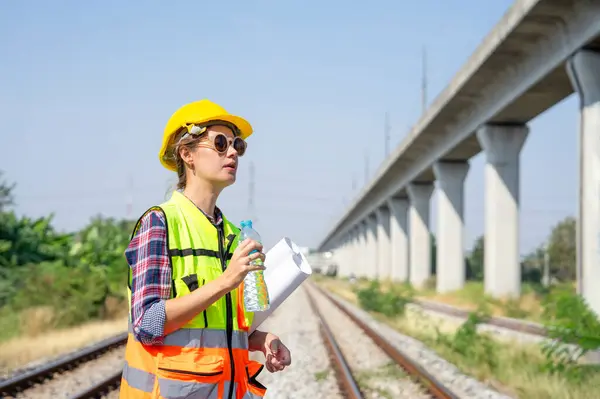 railway engineer or construction worker in hardhat and safety vest,holding paperwork is thirsty in the harsh sunlight,attractive caucasian female wears sunglasses drinking water while working outside