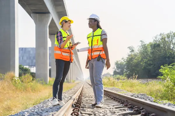 multi ethnic railway construction workers working outdoors,standing at rail tracks,discuss work,african woman technician having walkie talkie and blue print,caucasian female holding digital tablet