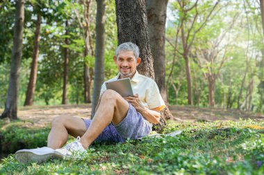 Mutlu Asyalı kıdemli bir adam ulusal parktaki bir ağacın altında dinleniyor, dijital tablet kullanarak aile ile iletişim kuruyor, modern yaşam tarzı, teknoloji, seyahat, rahatlama...